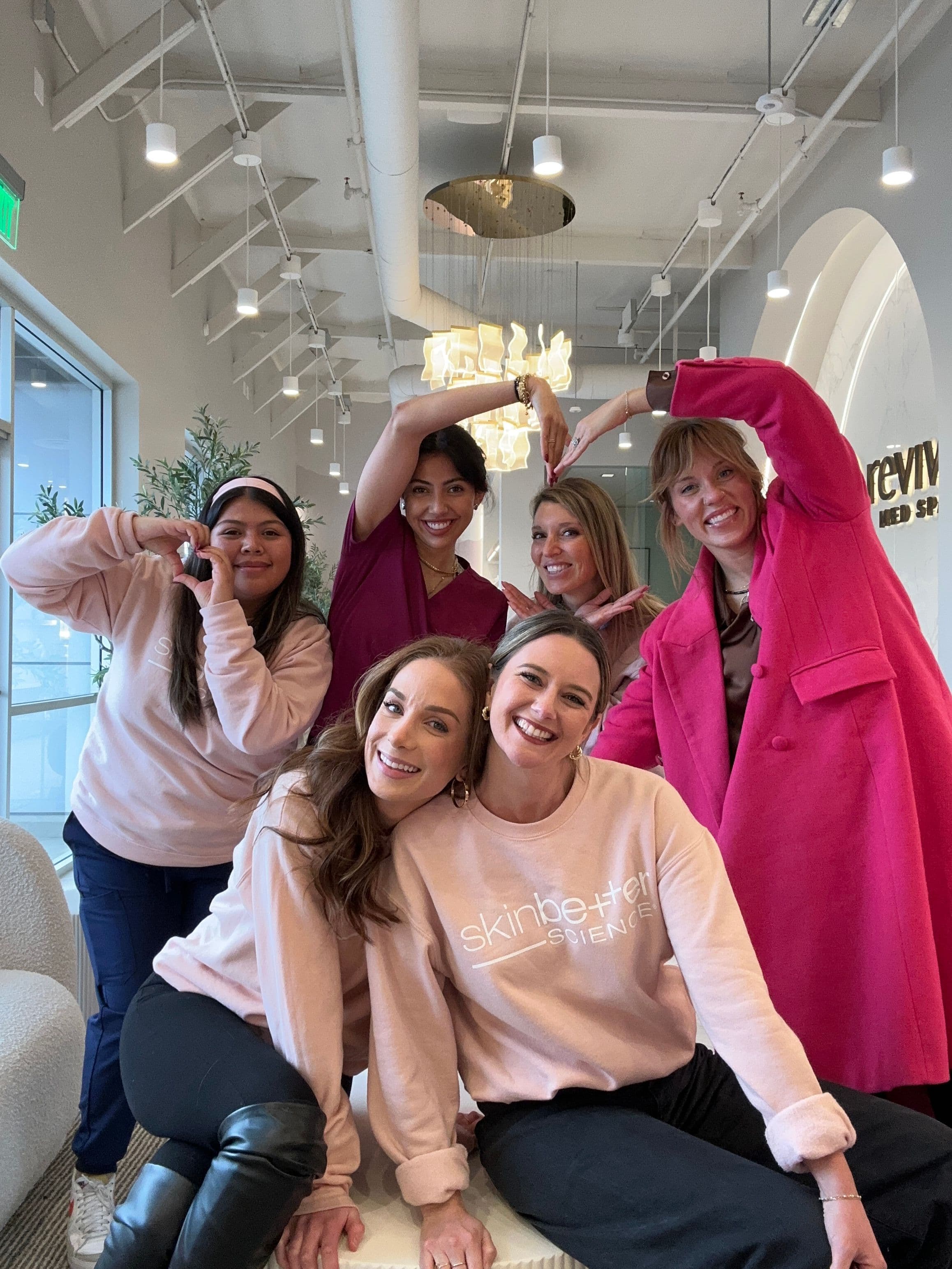 6 women dressed in pink smiling and forming hearts with their arms